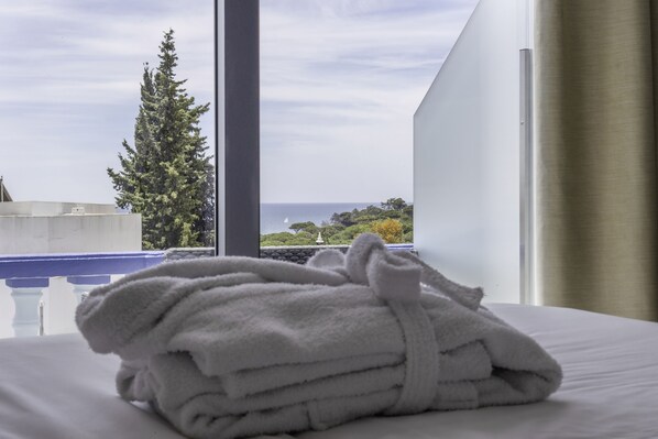 Una toalla blanca doblada con cuidado sobre una cama, con vista al mar y un árbol a través de la ventana.