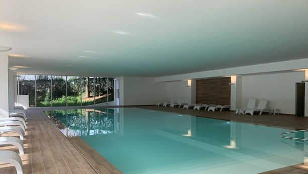 Une piscine intérieure moderne avec des parois en verre transparent, un plancher en bois et des chaises longues blanches disposées le long des côtés.