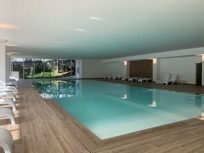 Een modern binnenzwembad met glazen wanden, houten vloeren en witte lounge fauteuils langs de randen.