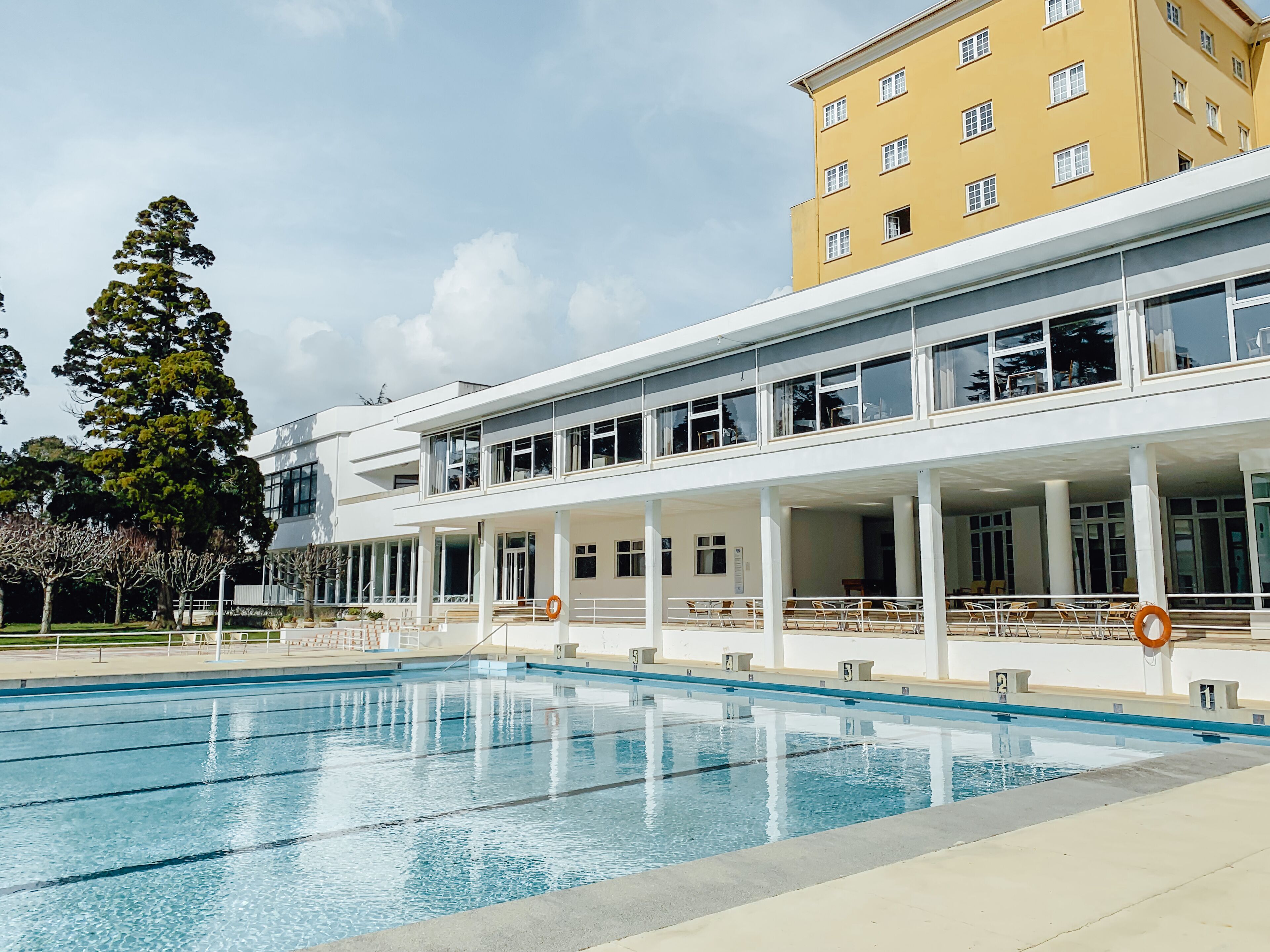 En moderne bygning med poolområde, der inkluderer en stor udendørs pool, flere siddepladser og en gul bygning i baggrunden.