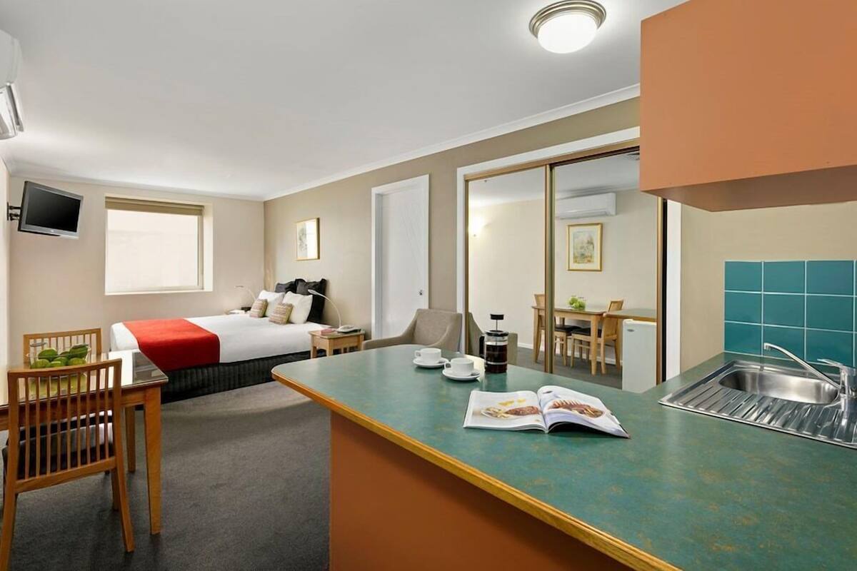 A compact hotel room with a bed, a small dining area, a kitchenette, and a TV mounted on the wall.