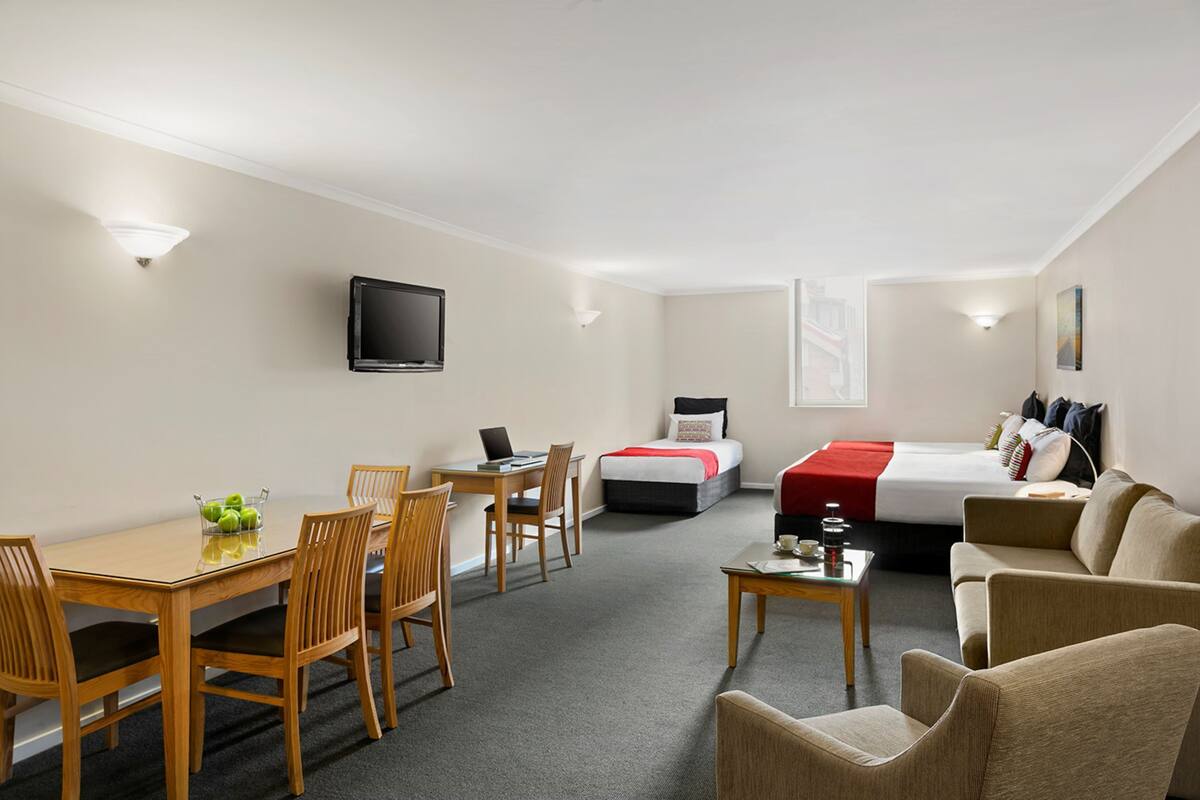 A hotel room with a bed, a sofa, a dining table with chairs, a TV, and a small table with a lamp.