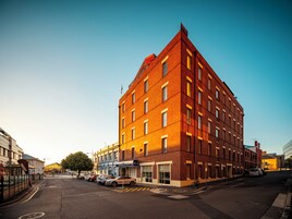 Un edificio de varios pisos con entrada en esquina, estacionamiento y estructuras más pequeñas adyacentes.