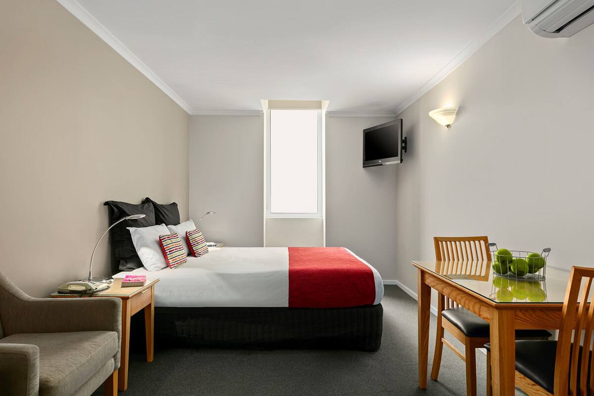 A hotel room with a bed, a desk with a chair, a TV mounted on the wall, and a window providing natural light.