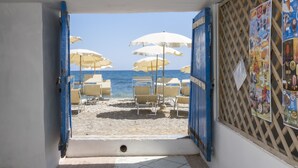 Ein offenes Durchgang zu einem Strandbereich mit Sonnenschirmen und Liegen, Blick auf das Meer, und eine Wand mit einem Poster.