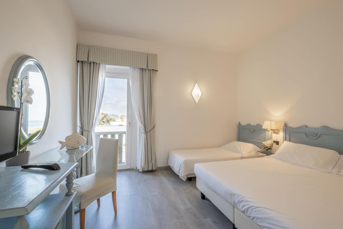 A hotel room with a desk, two beds, a chair, a window with curtains, a wall-mounted light, and a view of the outdoors.