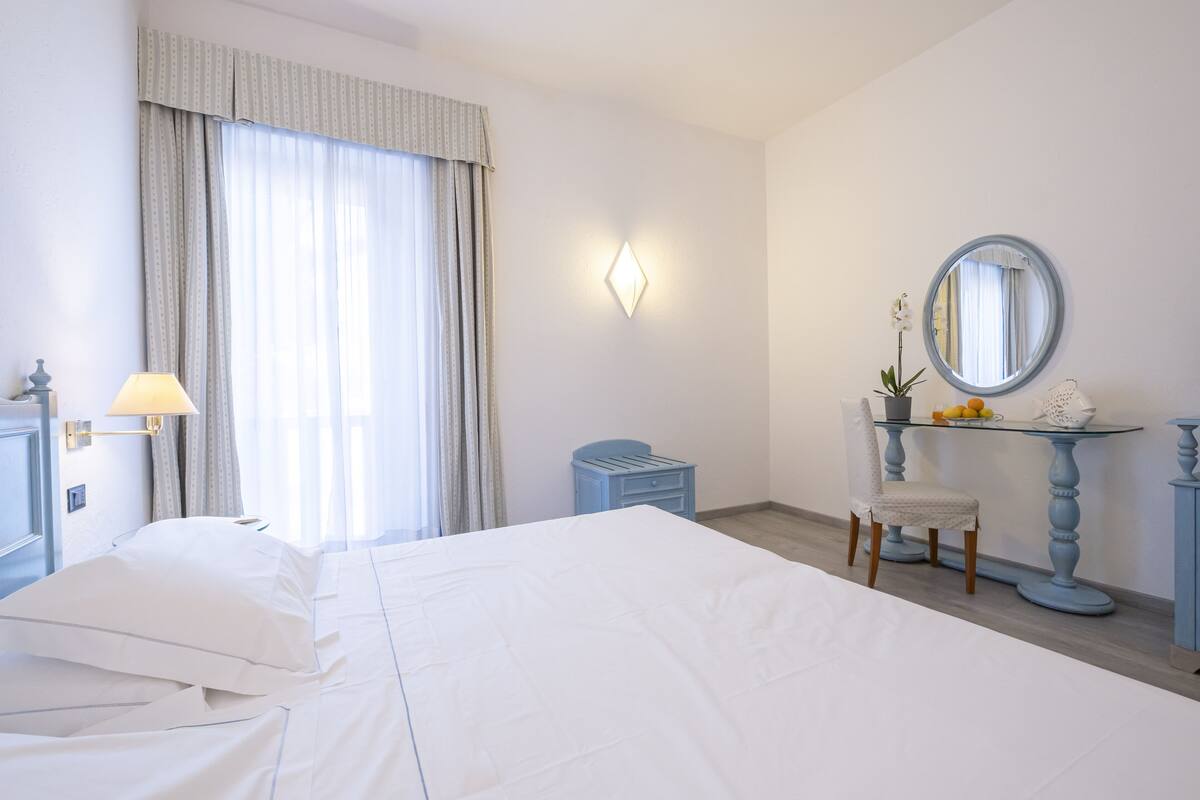 A hotel room with a bed, a desk with a chair, a mirror, and a small table with a vase of flowers.