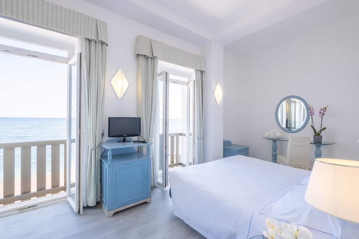A hotel room with a bed, a TV, a desk, and a view of the sea.