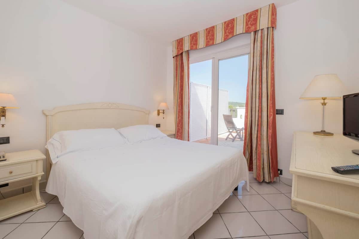 A hotel room with a bed, bedside lamps, a desk with a lamp, and a view of the outdoors.