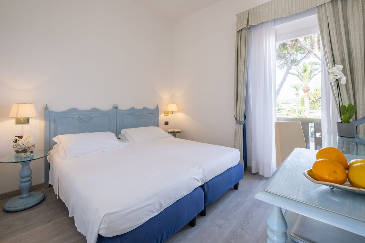 A bedroom with a bed, two bedside tables with lamps, a window with curtains, and a table with fruit.