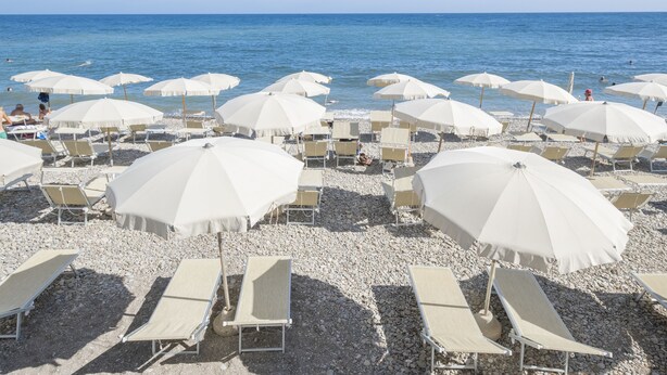 Ein Strand mit vielen weißen Sonnenschirmen und Liegestühlen auf Kies.