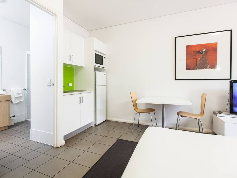 Une chambre d’hôtel moderne équipée d’un lit, d’un bureau, de deux chaises, d’un four à micro-ondes, d’un réfrigérateur et d’un tableau encadré au mur.