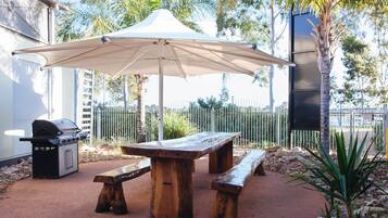An outdoor patio with a wooden table and benches, a barbecue grill, and a large umbrella.