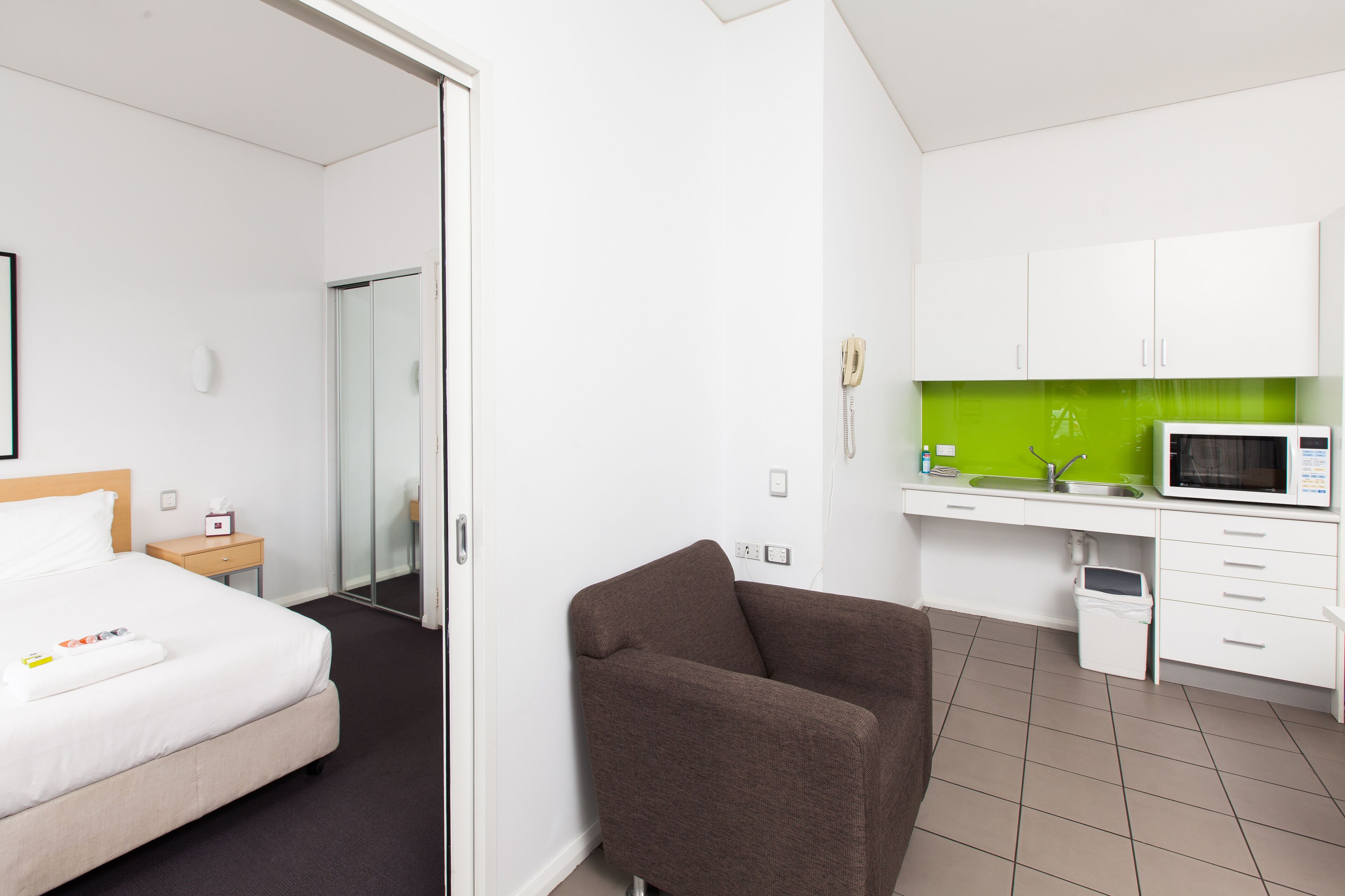 A hotel room with a bed, a brown armchair, a kitchenette with a microwave, and a glass door leading to a bathroom.