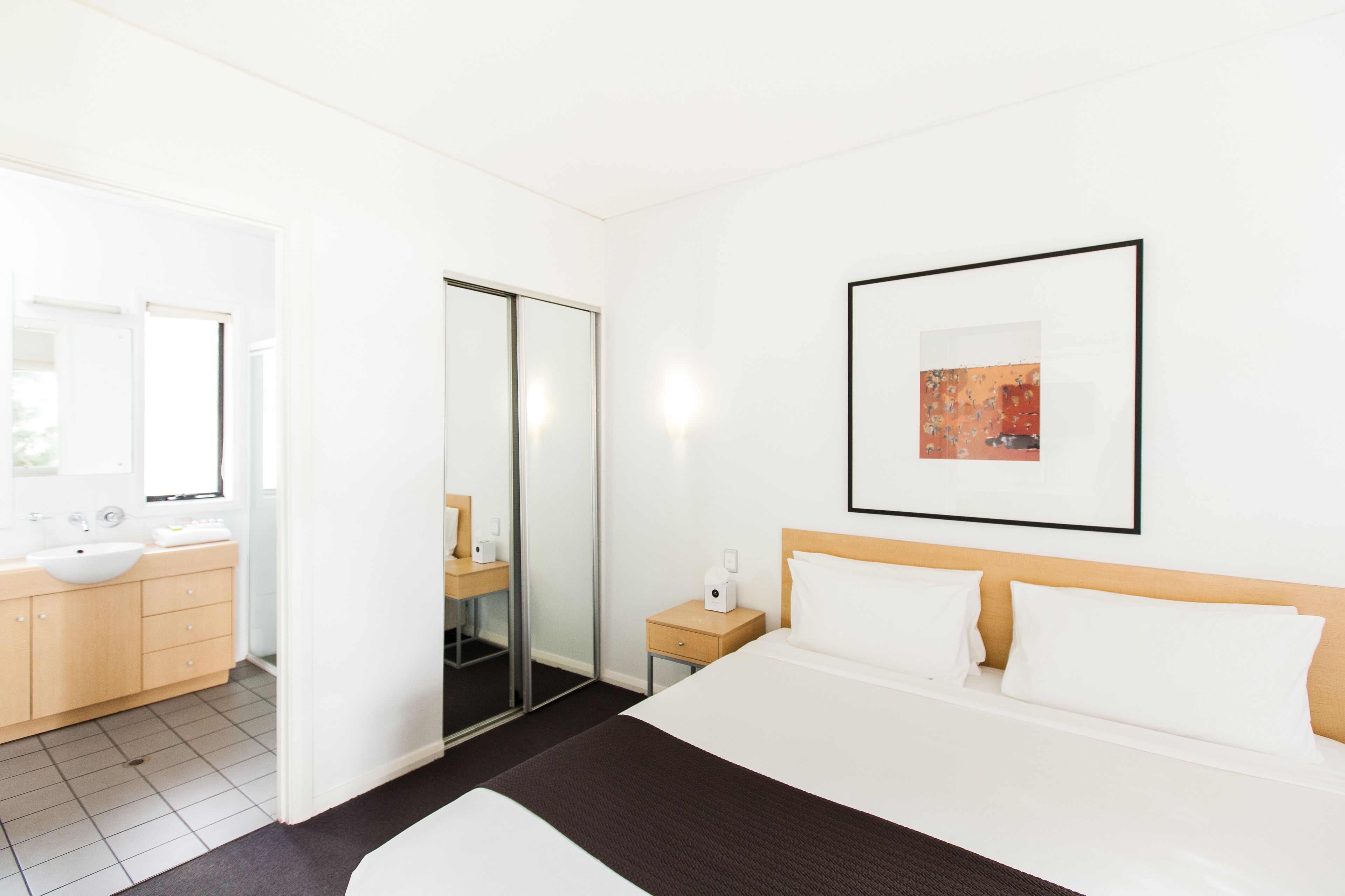 A hotel room with a bed, a bedside table, a nightstand, a mirror, a bathroom with a sink and toilet, and a framed picture on the wall.