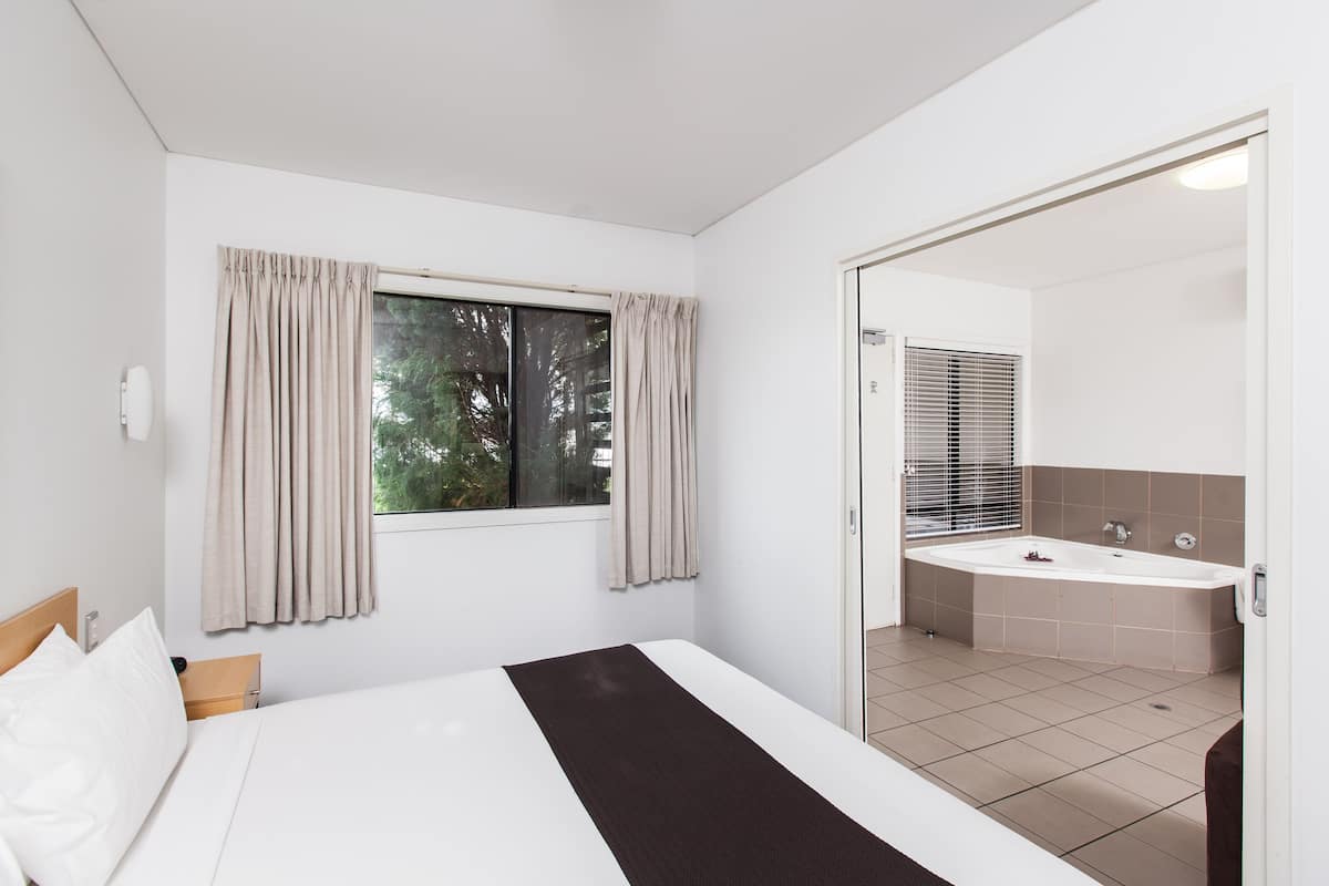 Ein Hotelzimmer mit Bett, Fenster mit Vorhängen sowie einem Badezimmer mit Waschbecken und Spiegel.