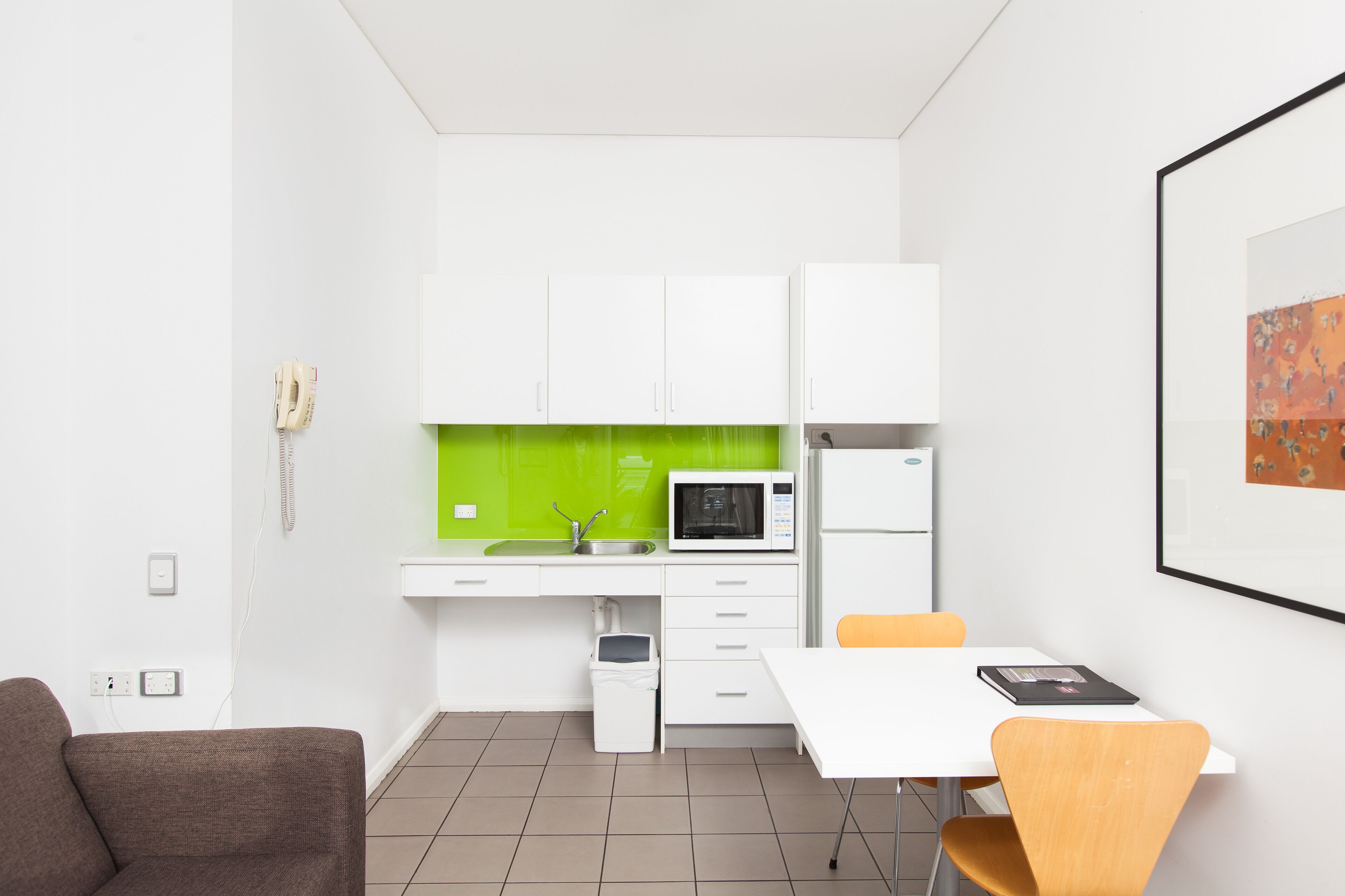 A compact kitchen with white cabinets, a microwave, and a sink. There is a small trash bin and a table with chairs.