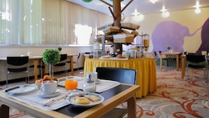 A breakfast setup with a fruit bowl, coffee, and orange juice on a table, with a decorative tree and a buffet in the background. - Hotel Santoli (Alto Reno Terme)