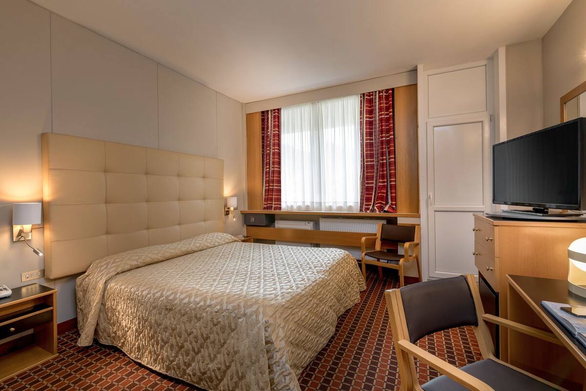 Une chambre d’hôtel avec un grand lit, un bureau avec une chaise, une télévision et une fenêtre avec des rideaux.