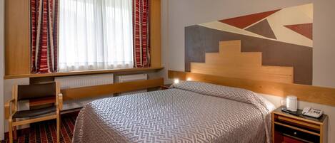 A hotel room with a bed, a desk, a chair, a radiator, a phone, and a window with curtains.