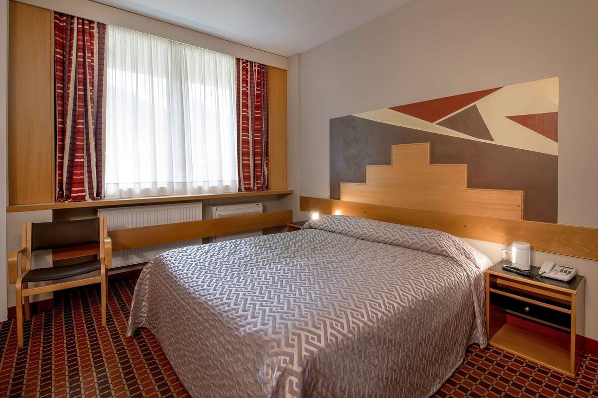 Une chambre d’hôtel équipée d’un lit, d’un bureau, d’une chaise, d’un radiateur, d’un téléphone et d’une œuvre d’art accrochée au mur.