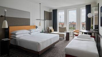 Habitación de hotel con una cama grande, dos camas más pequeñas, un escritorio, una silla, una mesa redonda y vistas a la ciudad.