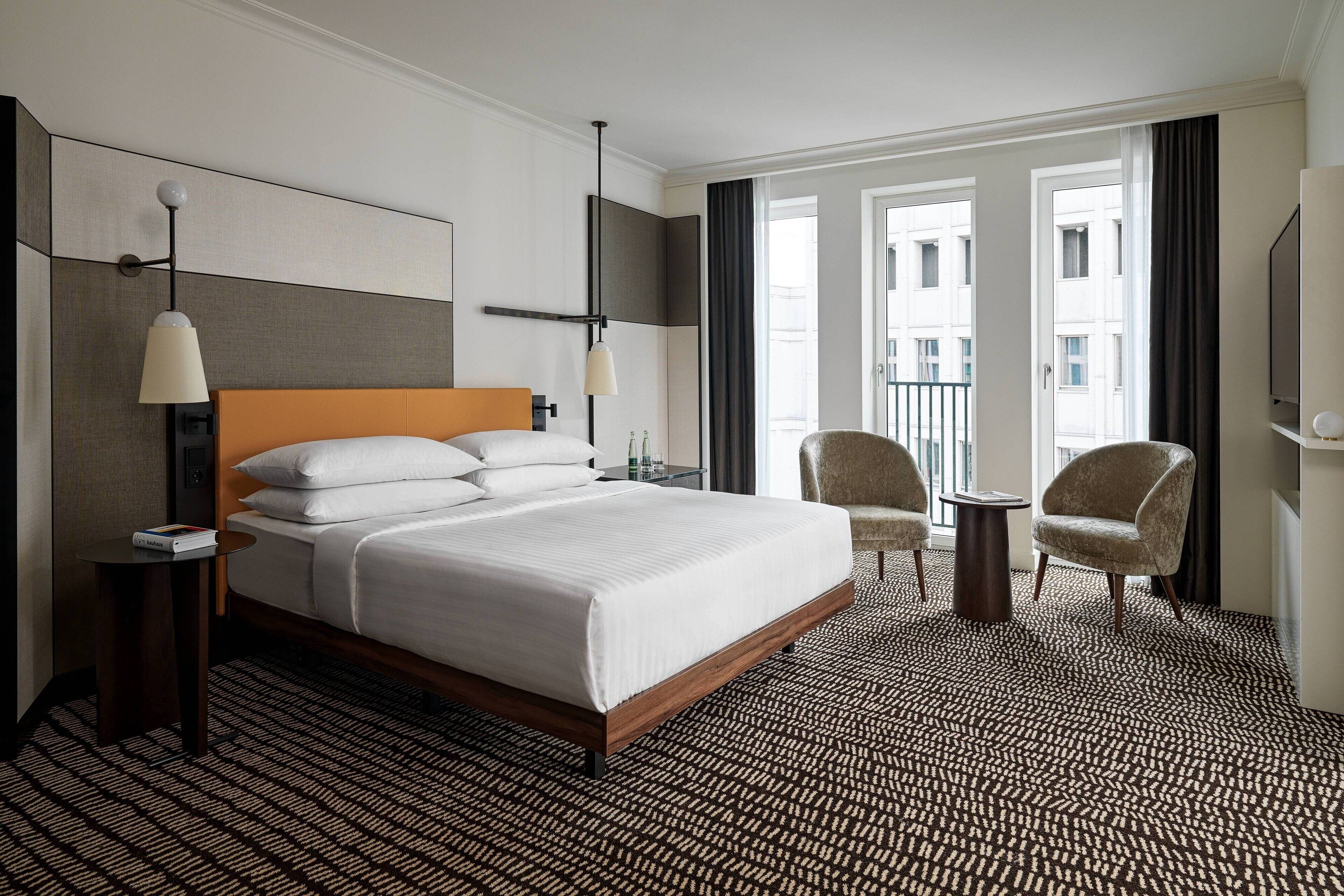 A hotel room with a large bed, two armchairs, a small table, a TV, and a view of the city through large windows.