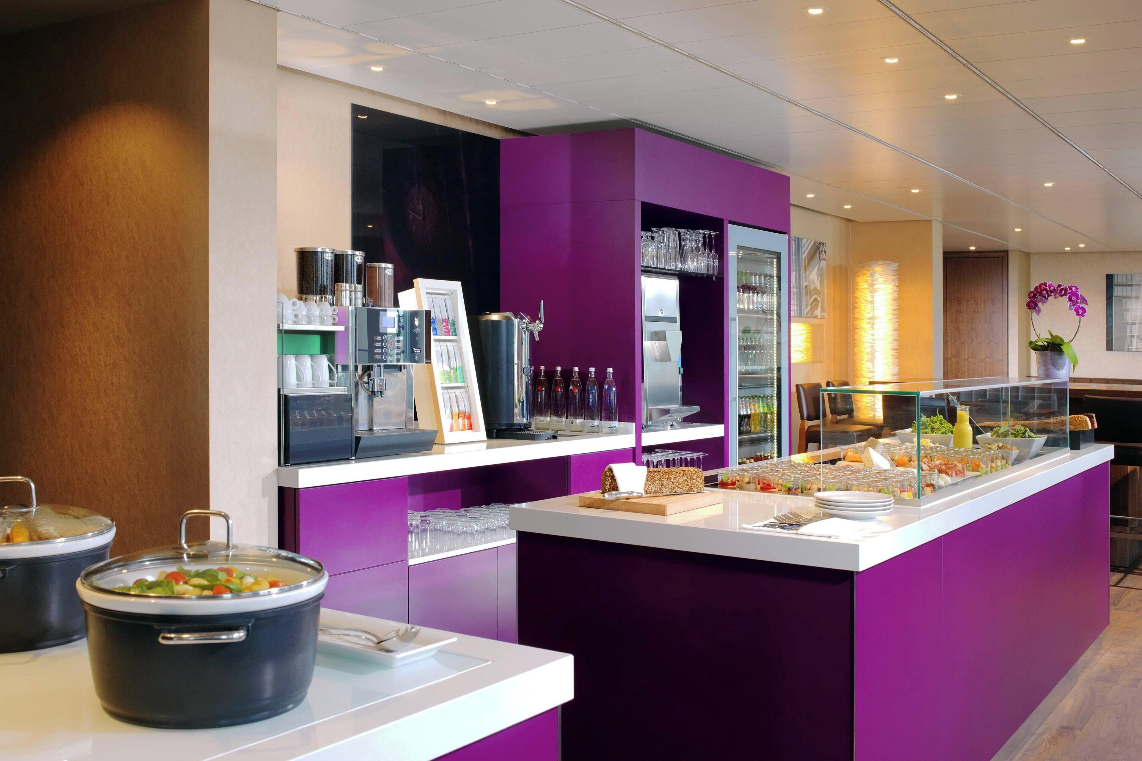 Una barra de desayuno moderna con una pared de acento púrpura, dispensadores de bebidas tipo autoservicio y diversas opciones de comida.