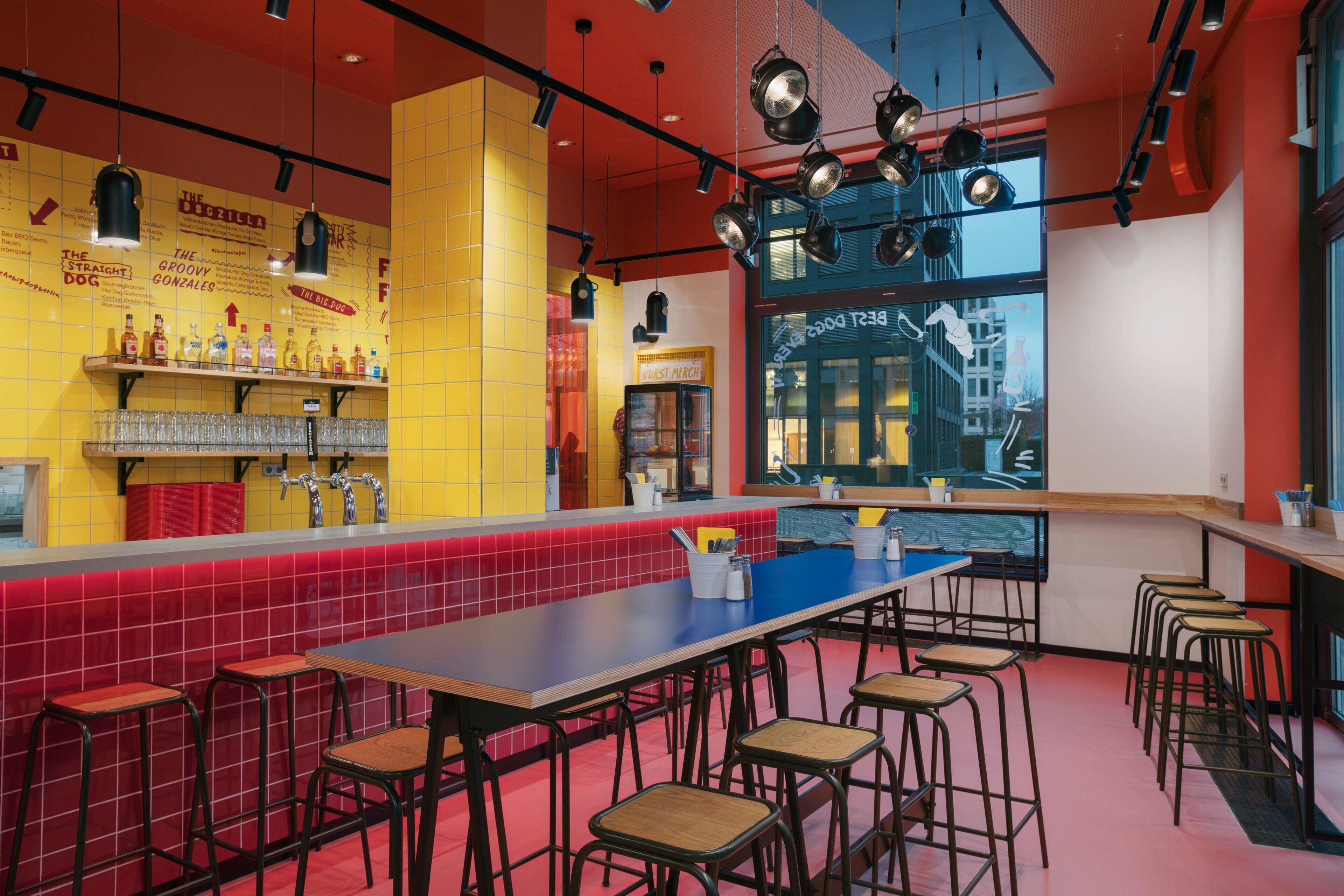 A vibrant bar area with a red-tiled bar counter, multiple stools, and hanging lights.