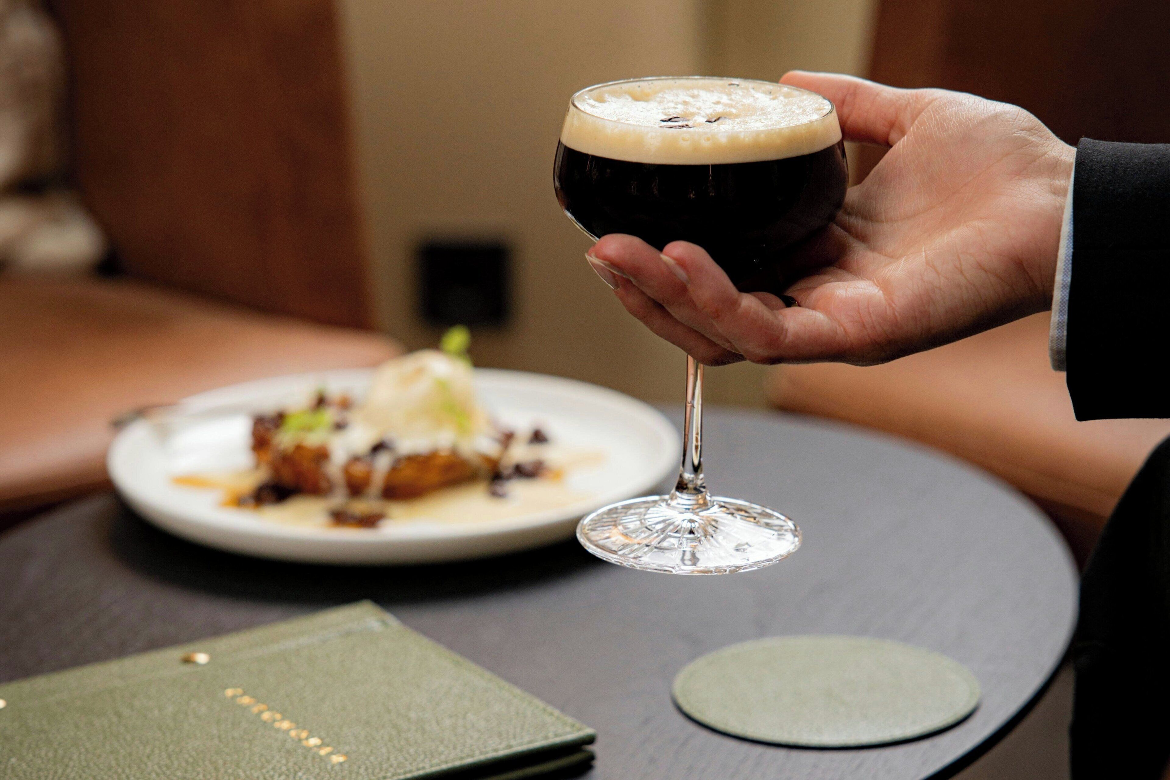 Una mano sosteniendo una jarra de cerveza oscura con espuma, un plato de comida y un menú verde sobre una mesa redonda.