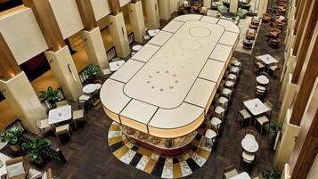 A spacious indoor area with a central table and multiple seating arrangements, surrounded by columns and potted plants.