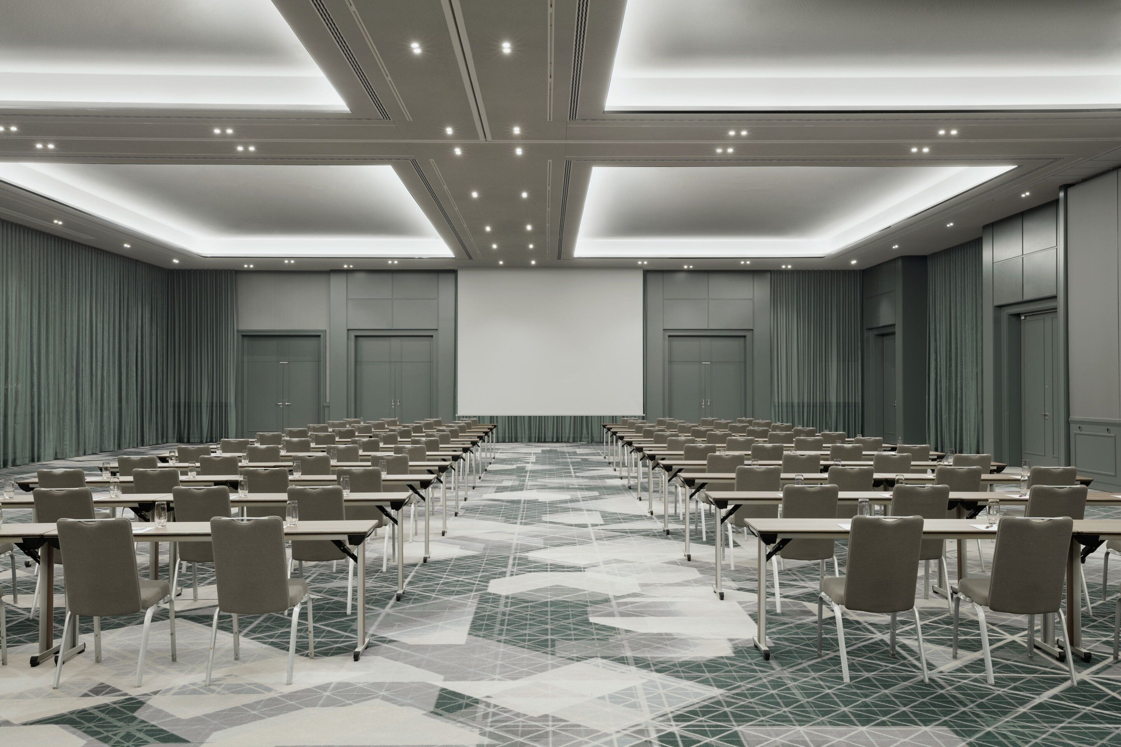 Un salón de conferencias con filas de mesas y sillas, una pantalla grande y un suelo con diseño.