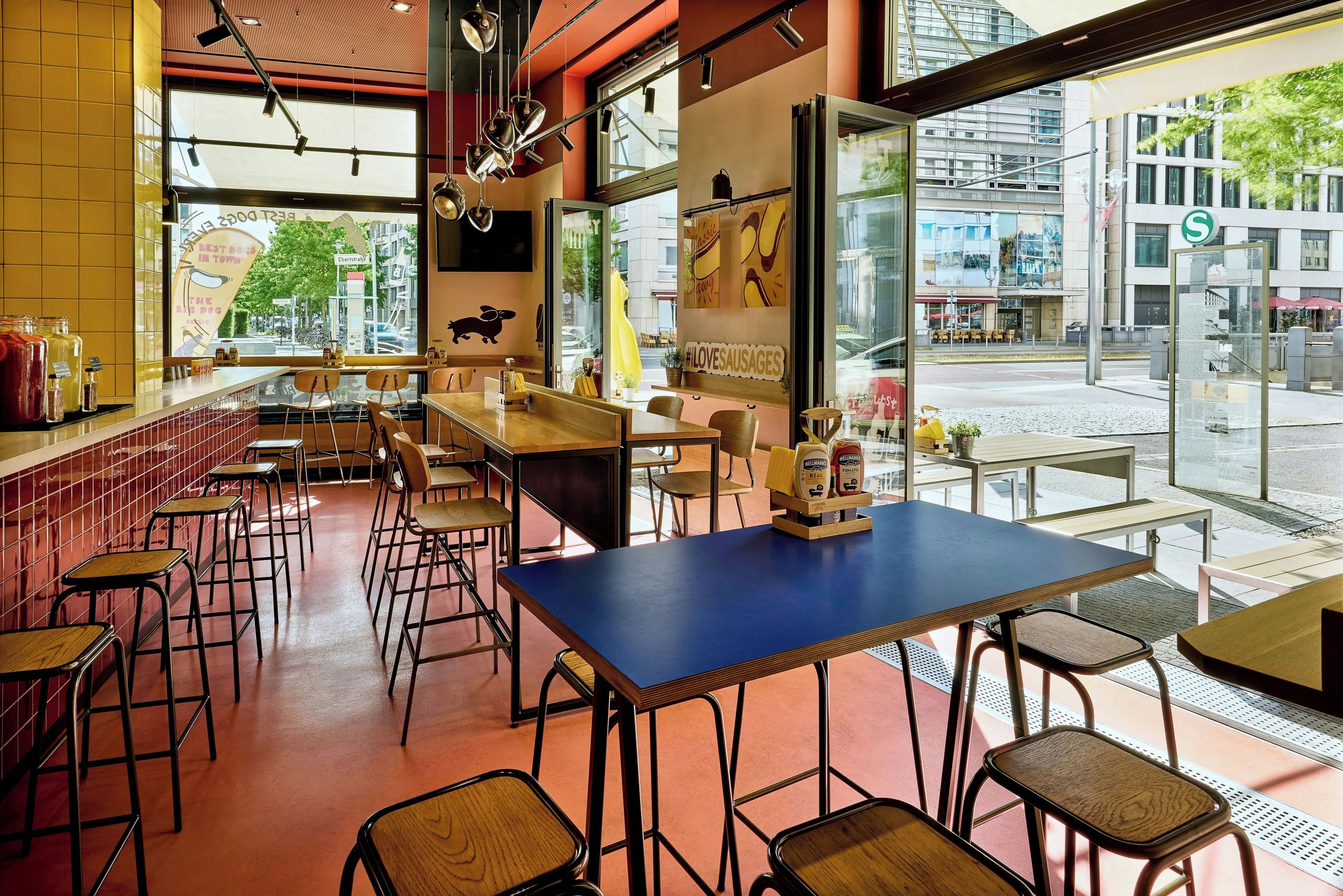 A restaurant interior with multiple tables and chairs, a large window with a view of the street, and a sign on the wall.