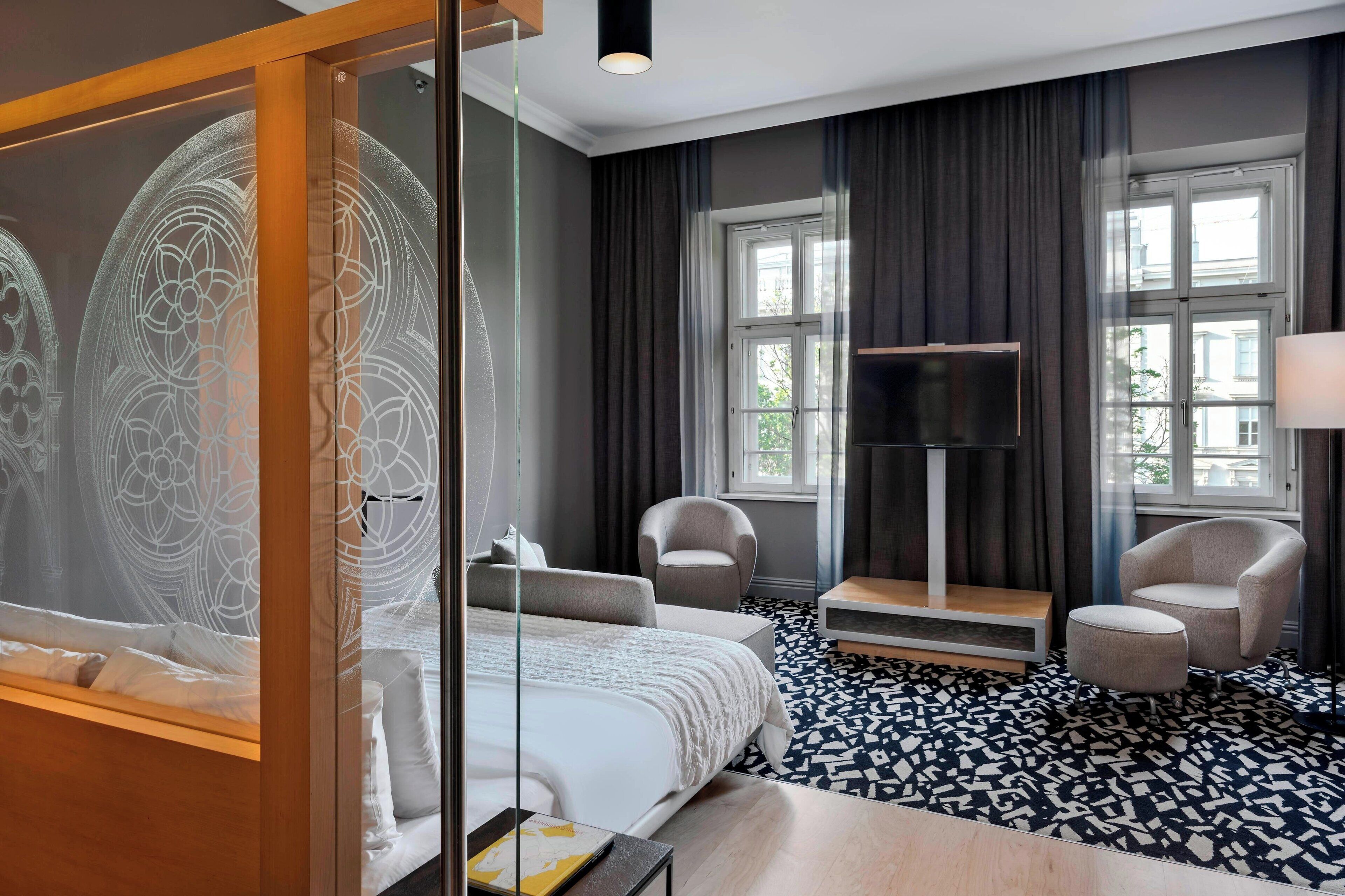 A modern hotel room with a large bed, two armchairs, a TV, and a patterned floor.