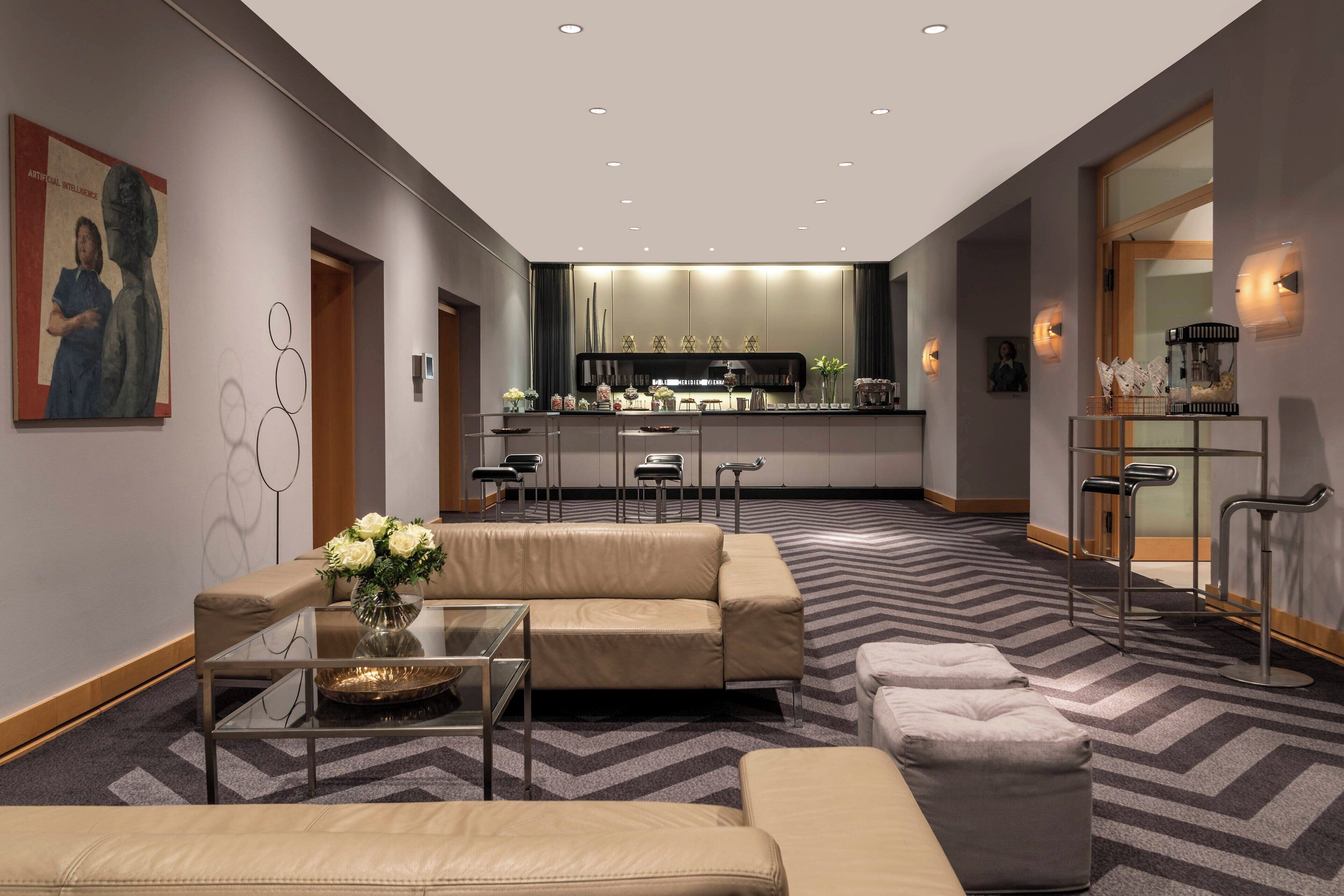 A modern hotel lobby with a central seating area, a kitchen in the background, and a bar area with stools.