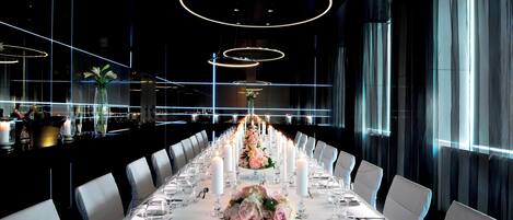 Een lange eettafel, gedekt met witte linnen, kaarsen en bloemstukken, in een modern restaurant met een weids stadsgezicht.