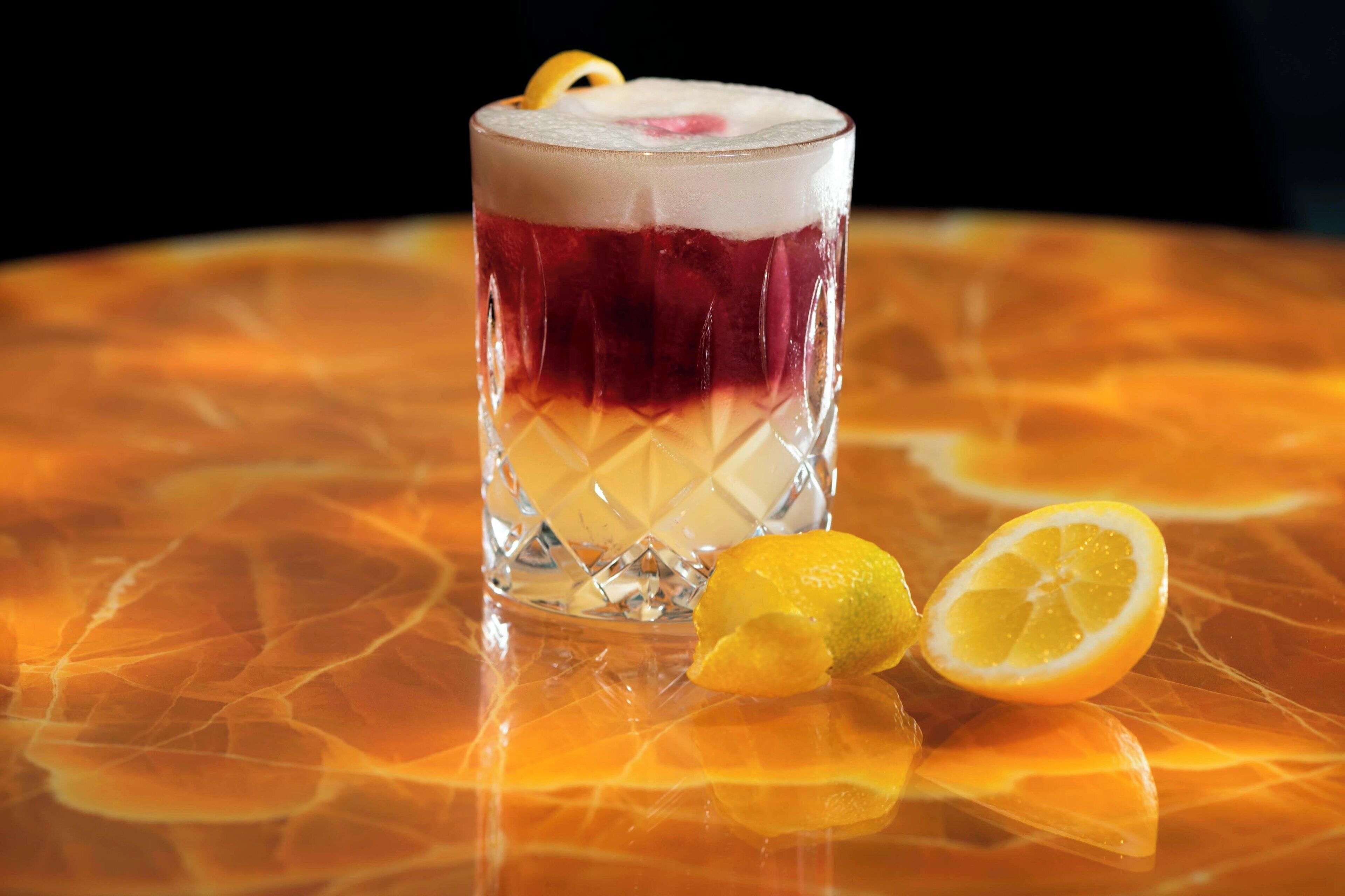 A glass with layered drinks and a lemon slice on a reflective orange surface.