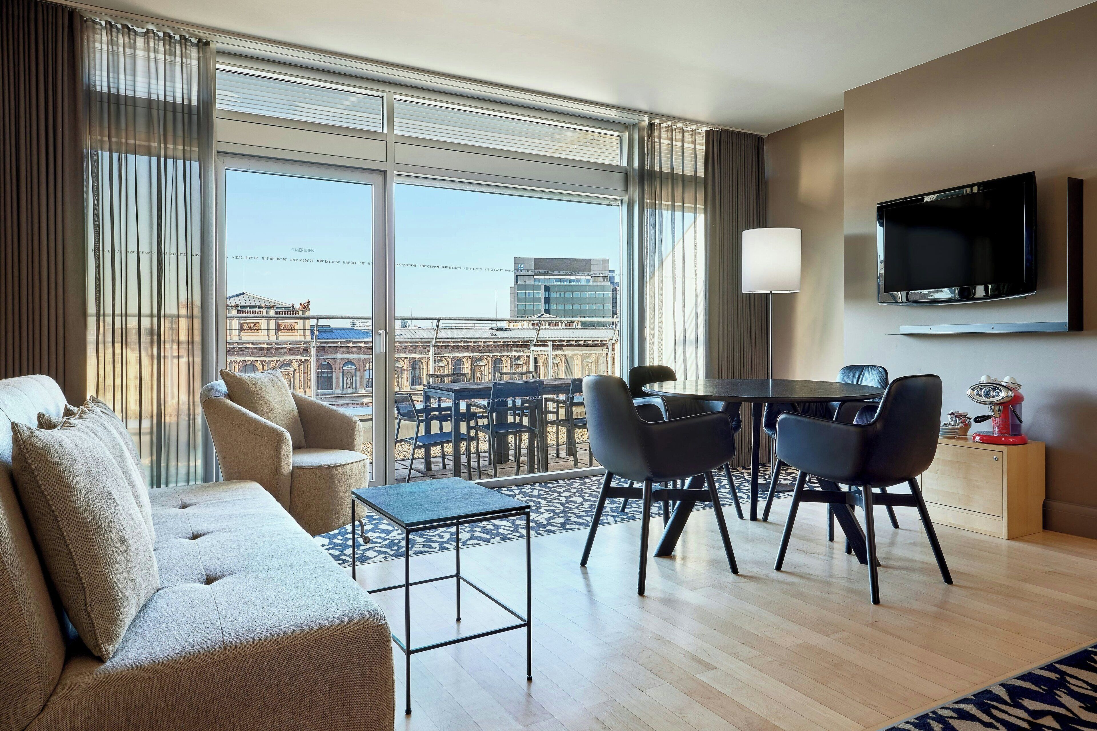 A modern hotel room with a large window, a sofa, a dining table with chairs, a TV mounted on the wall, and a lamp.