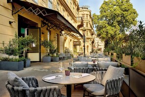 Un café al aire libre con mesas redondas y sillas tapizadas en gris, rodeado de plantas en macetas y un edificio histórico al fondo.