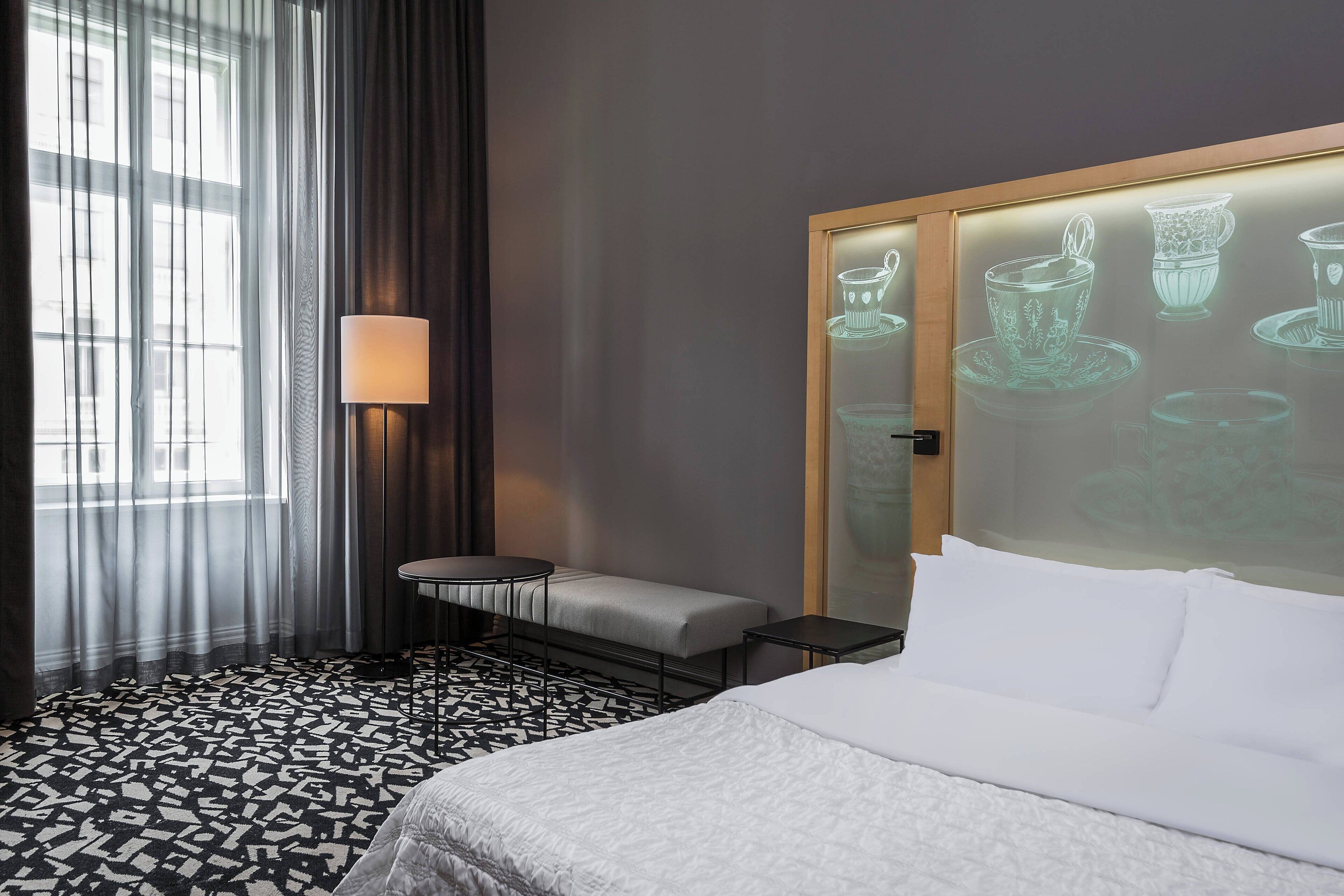 Een hotelkamer met een bed, een tafeltje, een bankje, een lamp en een muur waarop kopjes worden geprojecteerd.