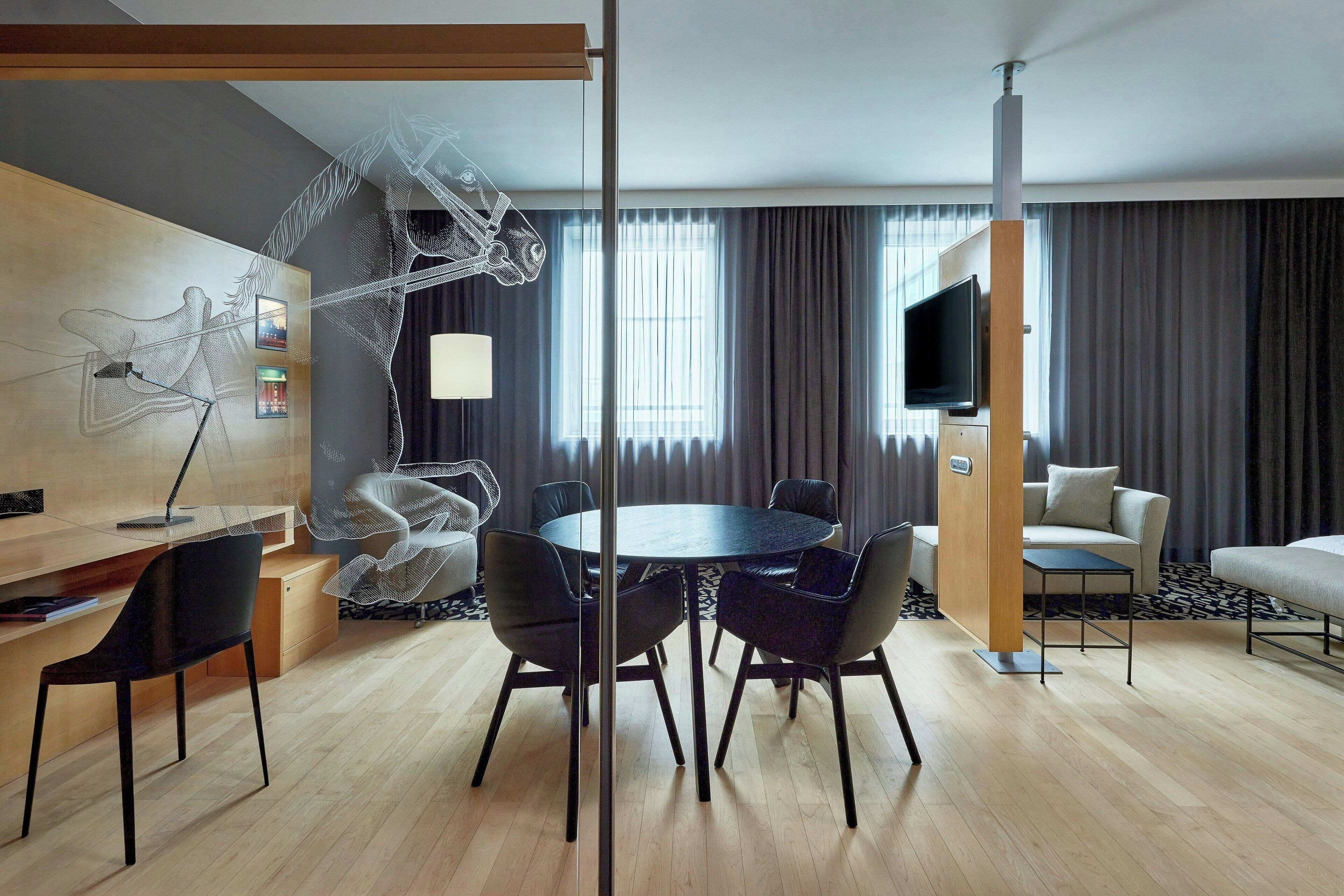 Una habitación de hotel moderna con una mesa de comedor redonda, sillas negras, un sofá, un televisor y un ventanal con cortinas.