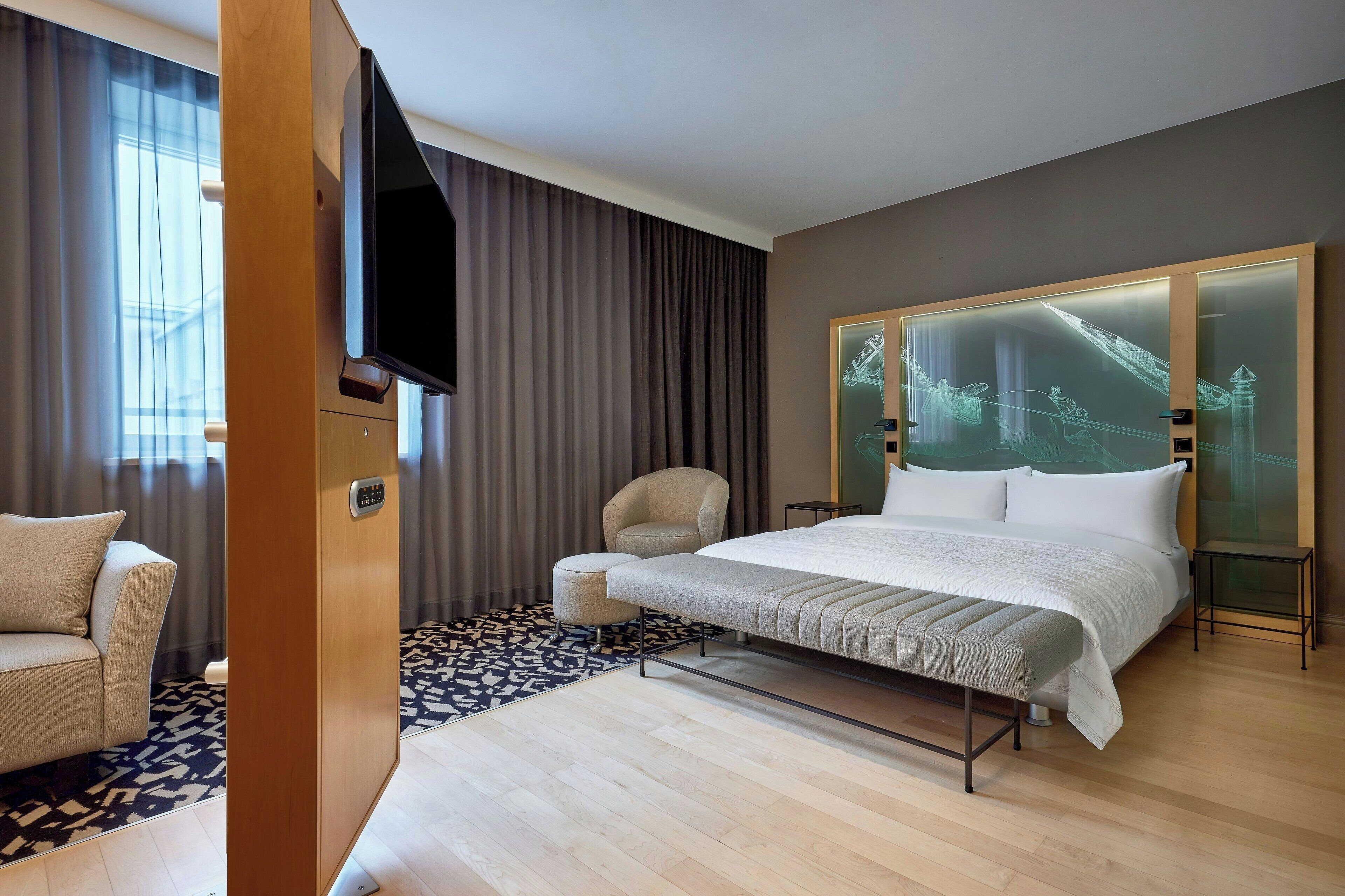 A modern hotel room with a large bed, a flat-screen TV mounted on the wall, a comfortable armchair, and a patterned rug.