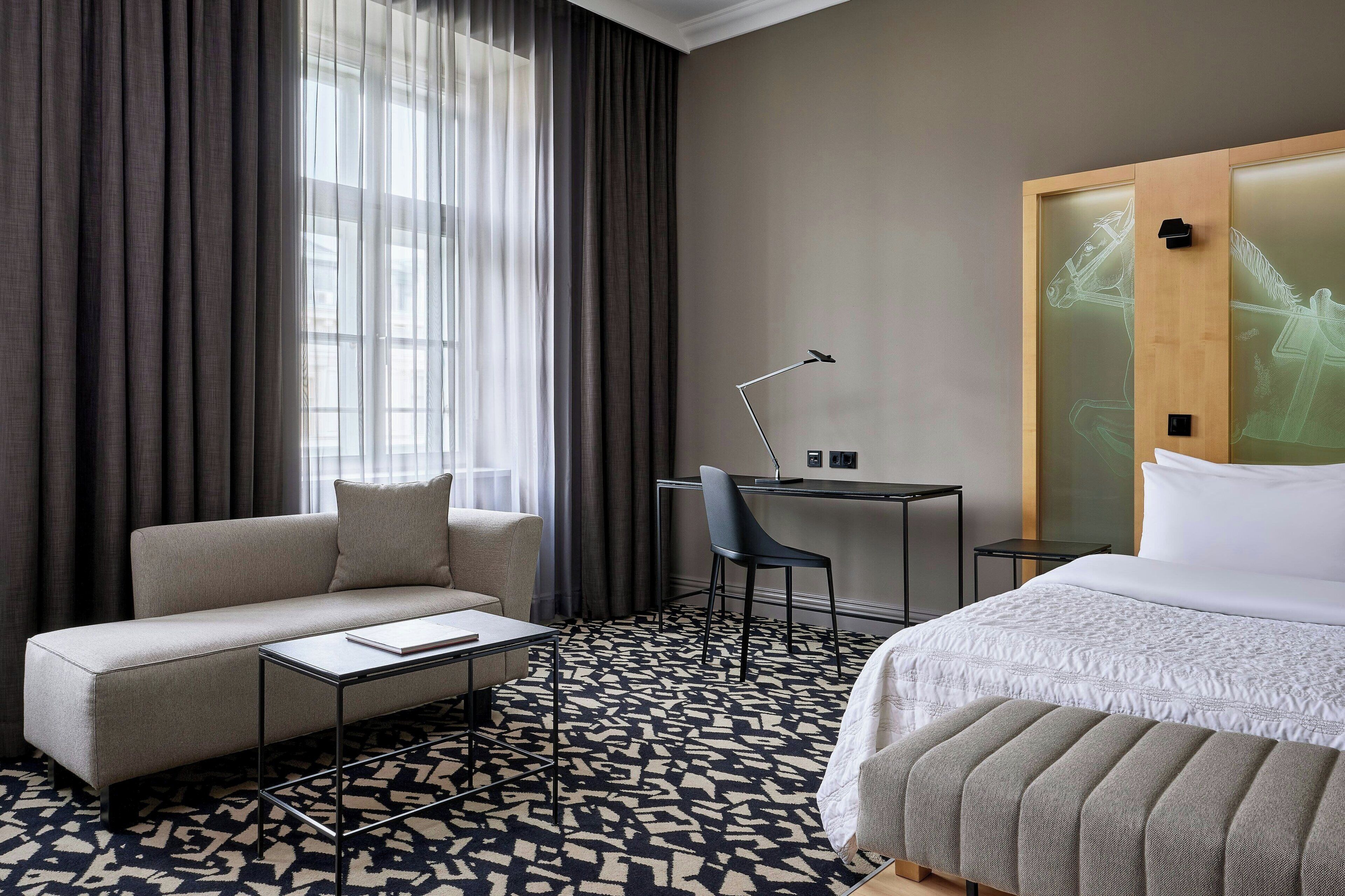 A hotel room with a sofa, a small table, a chair, a bed, and a desk with a lamp.