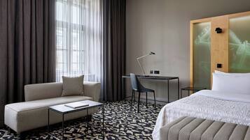 A hotel room with a sofa, a small table, a chair, a bed, and a desk with a lamp.