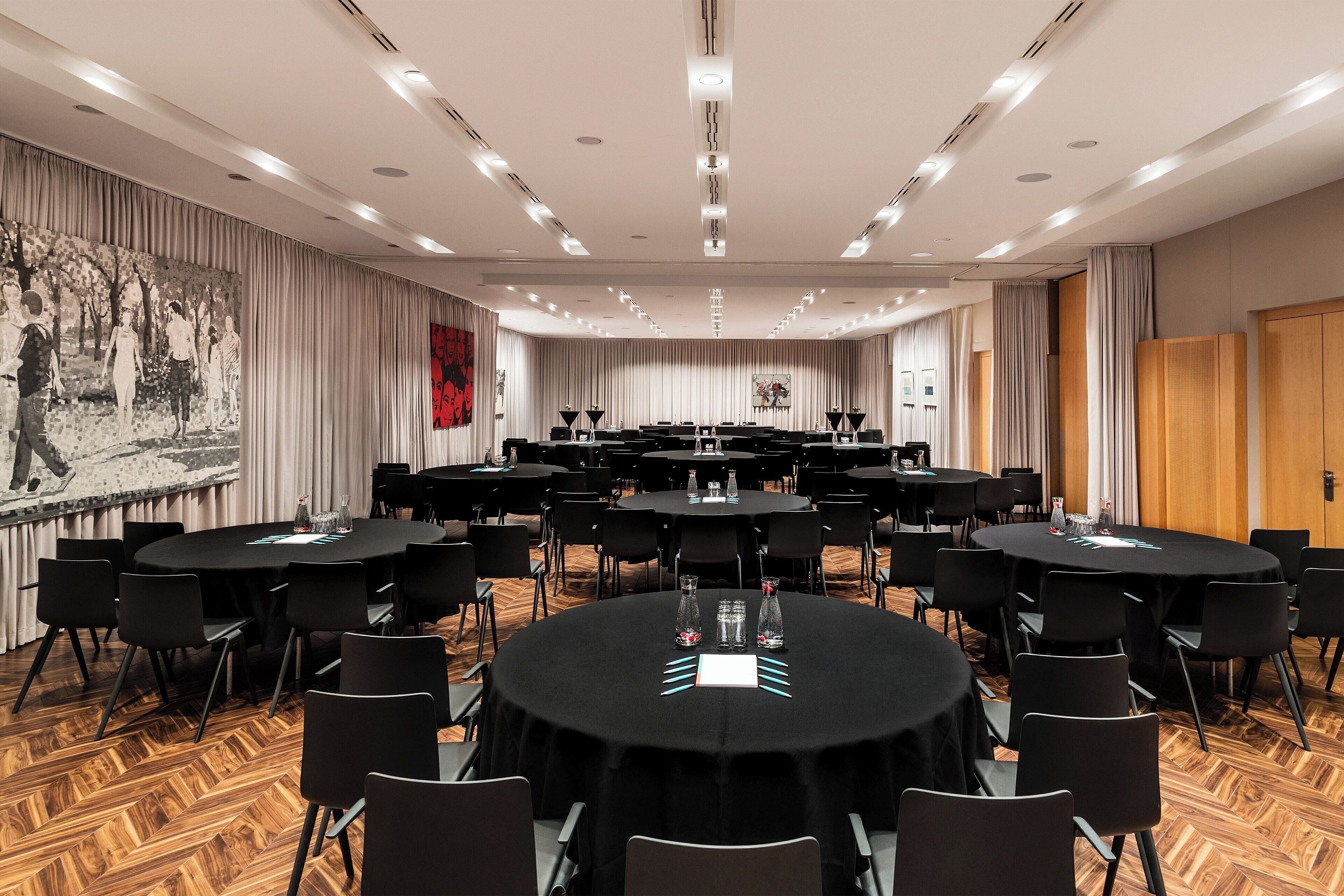 Uma sala de conferências com várias mesas redondas, cadeiras pretas e piso de madeira em padrão herringbone.
