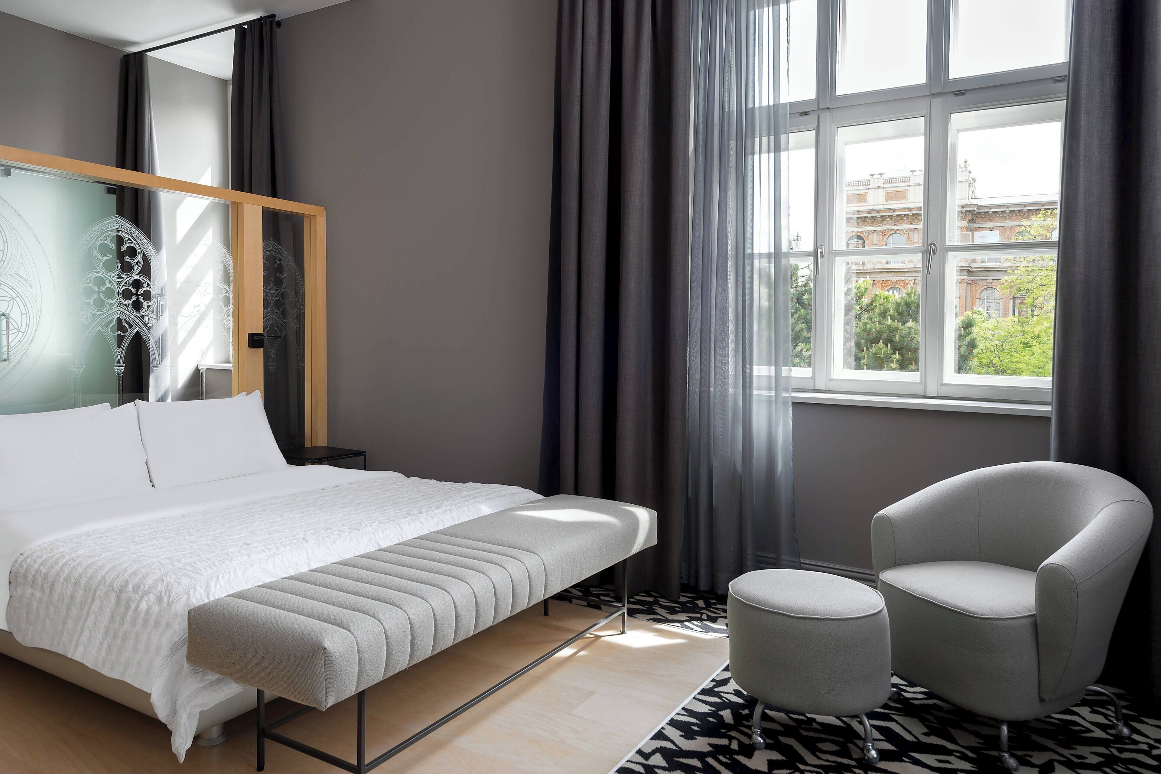 A modern hotel room with a large bed, a small armchair, a round ottoman, and a window with a view of buildings.