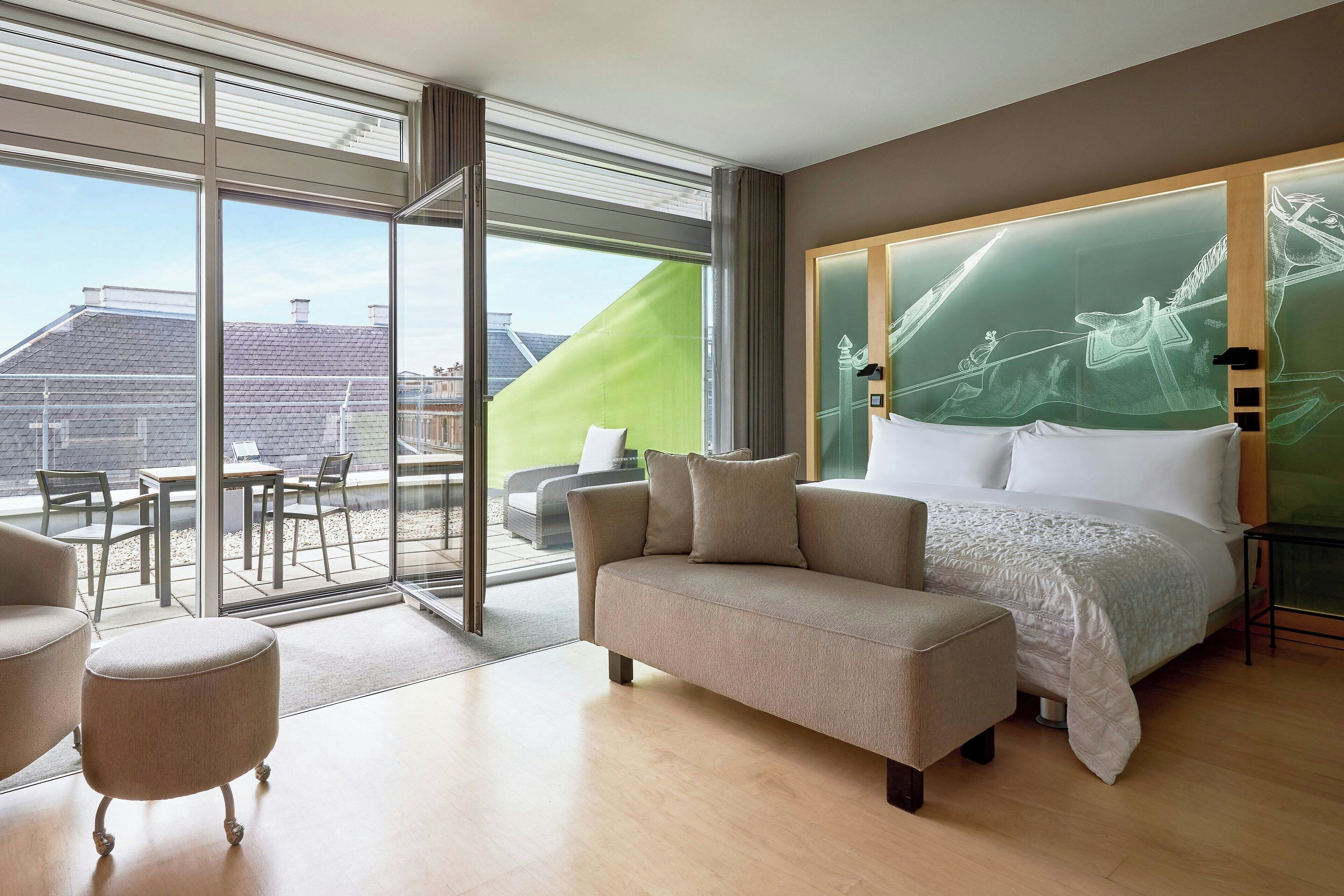 A modern hotel room with a large bed, a sofa, a small ottoman, and a balcony with outdoor furniture.