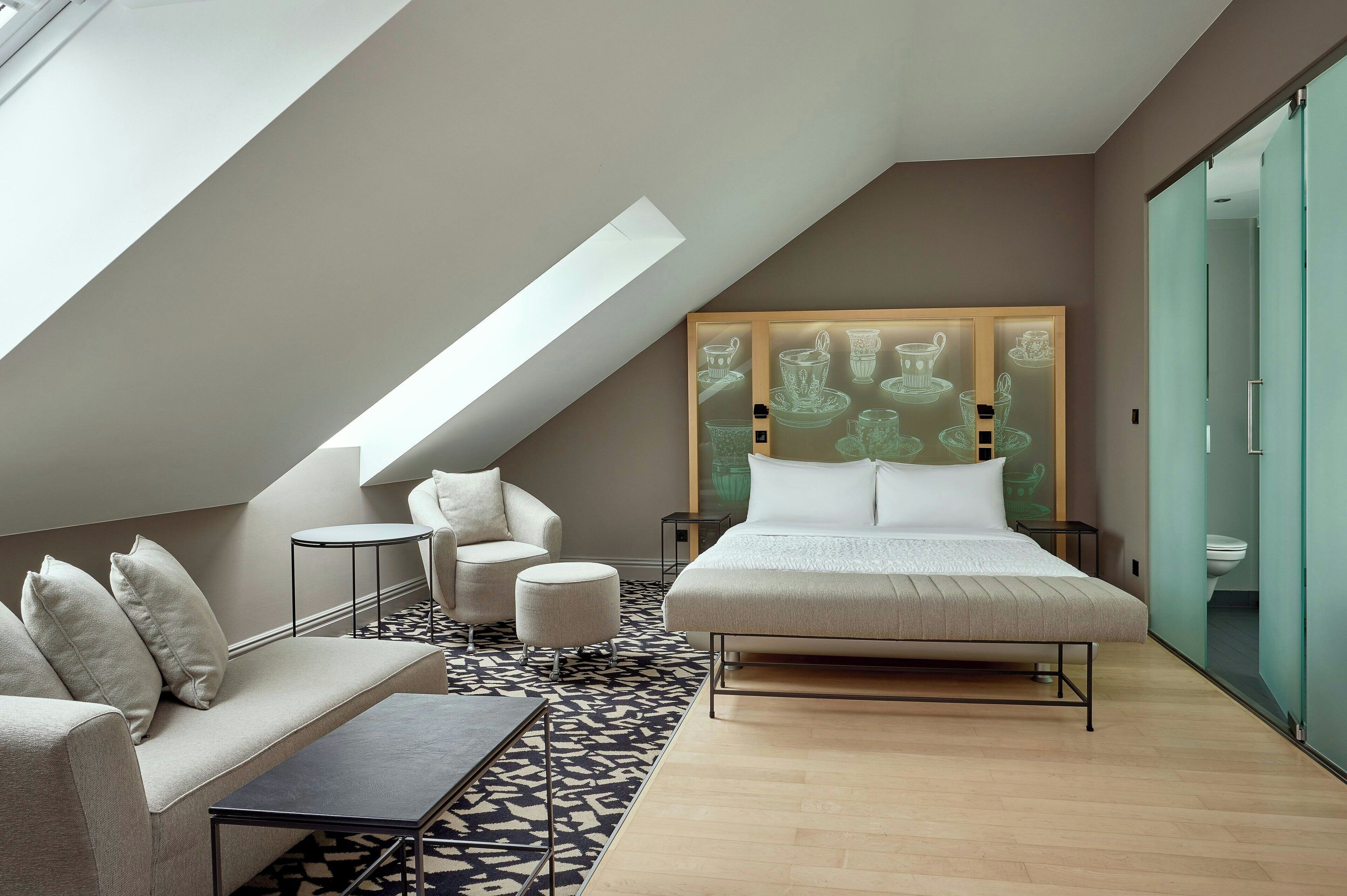 Een moderne slaapkamer met een schuine kap, een bed, een bank, een klein tafeltje en een kamerkleed met een patroon.