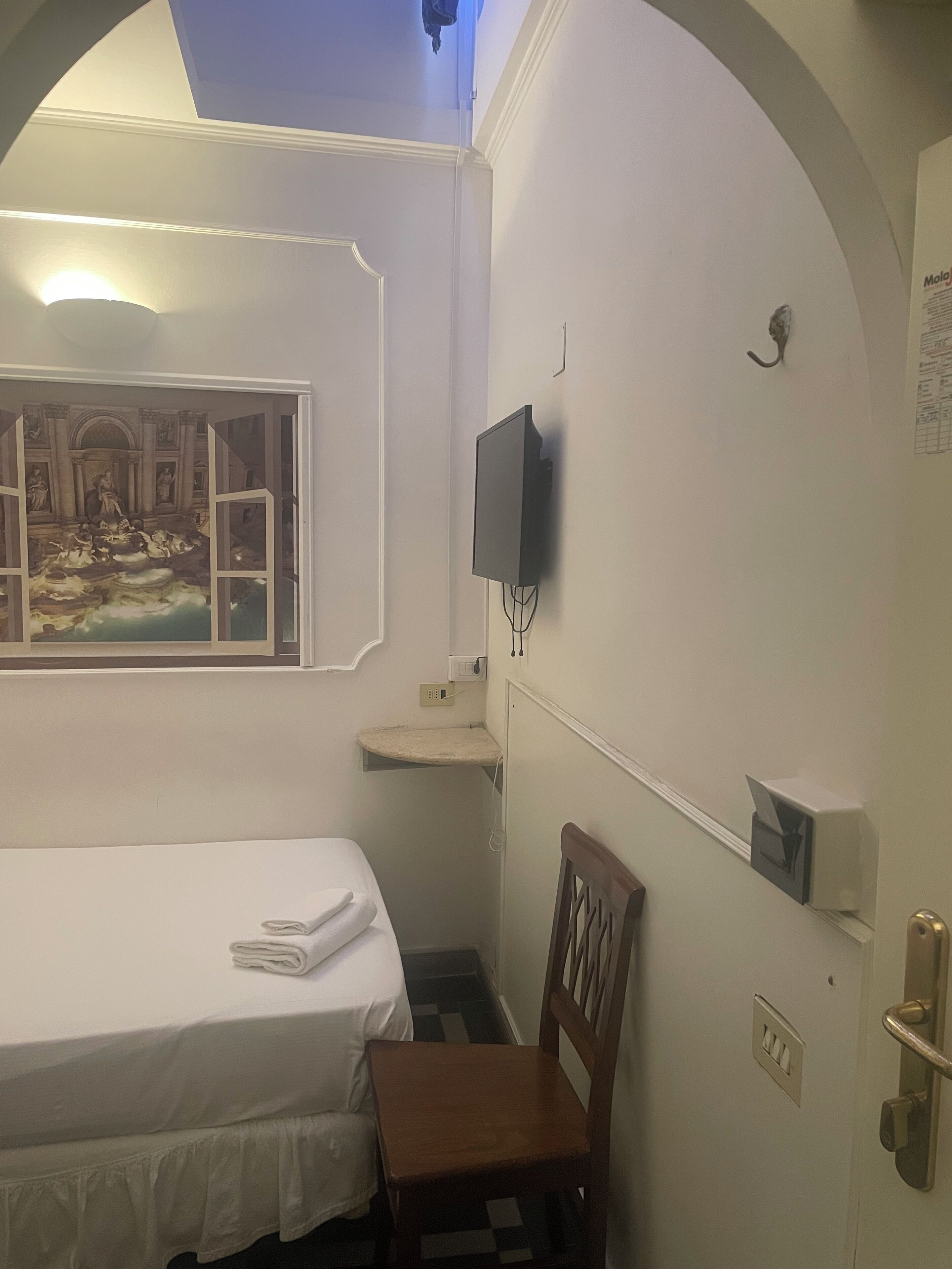Habitación de hotel con cama, una silla, televisor montado en la pared y un estante pequeño.