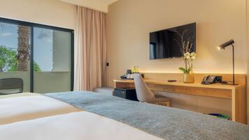 A hotel room with a large bed, a desk with a computer, a chair, a TV mounted on the wall, and a window with a view of trees.